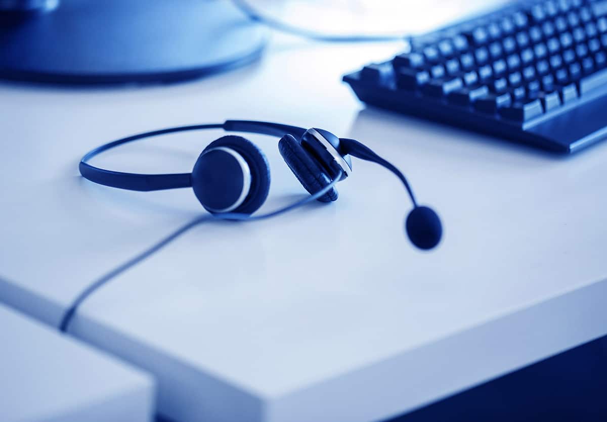 A pair of headphone with a microphone lay on a white desk with a computer in the background