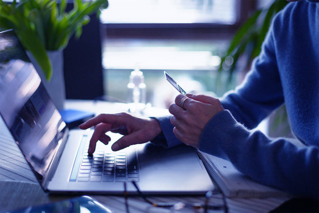 hands of a man sitting at a desk hold a credit card with one and is pointing to a screen of an open laptop with the other